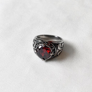 May include: A silver ring with a large, round, deep red gemstone. The ring has intricate, swirling patterns around the gemstone and on the band, giving it a vintage or gothic look. The ring is displayed on a white background.