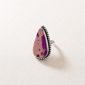 Peut inclure: Bague en argent avec une grosse pierre en forme de goutte d'eau. La pierre présente un mélange de teintes violettes et roses avec des mouchetures marron. La bague a une bordure perlée et un anneau simple.