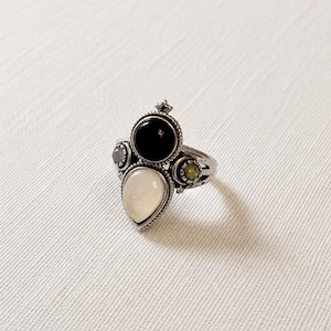 May include: A silver ring with a cluster of gemstones. The ring features a large, pear-shaped, off-white stone at the bottom, with a black circular stone at the top. Smaller grey and yellow-green stones are also present.