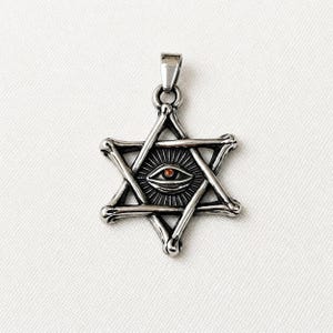 May include: A silver-colored pendant featuring a six-pointed star design with an eye in the center. The eye has a red center and is surrounded by sun rays. The pendant has a bail for attaching to a chain.