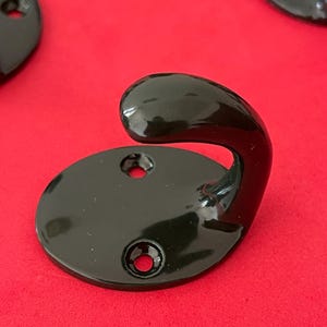 May include: Close-up of several black metal coat hooks. Each hook has a rounded base with two screw holes and a curved hook. The hooks are on a red background.