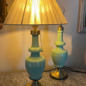 Pode incluir: Duas luminárias de mesa combinando bases de cerâmica azul clara e detalhes em metal dourado. Cada luminária tem uma cúpula plissada creme. As luminárias estão sobre uma superfície com efeito de mármore.