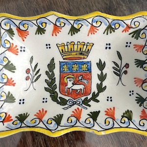 Könnte beinhalten: Eine weiße Keramik-Schale mit gelbem Rand und einem blauen, roten und grünen Blumendesign. In der Mitte der Schale befindet sich ein Wappen mit einer Krone, einem Schild und einem Lamm.