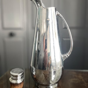 May include: A silver pitcher with a rounded body and a curved handle. The pitcher has a polished finish and a cork stopper.
