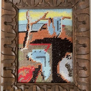 May include: A framed cross-stitch artwork featuring a surreal landscape with a brown, carved wooden frame. The artwork depicts a scene with blue, orange, and black elements, with a yellow and blue sky. The frame has a detailed, ornate design.