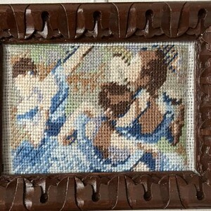 May include: A framed needlepoint artwork featuring a pastoral scene with figures in blue and brown hues. The artwork is encased in a dark brown, intricately carved wooden frame, adding a vintage aesthetic.