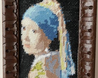 May include: A framed needlepoint of a woman with a pearl earring, reminiscent of Vermeer's painting. The artwork features a blue headscarf, a white collar, and a dark brown, ornate wooden frame. The needlepoint is a detailed cross-stitch.