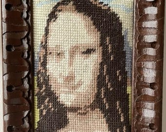 May include: A framed needlepoint portrait of a woman with long dark hair. The artwork is in a dark brown, carved wooden frame. The needlepoint uses shades of brown, beige, and blue to depict the subject's face and background.