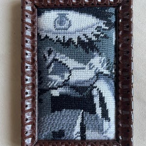 May include: A framed cross-stitch artwork depicting a grayscale landscape. The piece is set within a dark brown, decorative frame with a scalloped edge, giving it a vintage appearance.