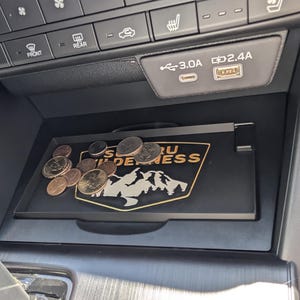 Pode incluir: Uma bandeja preta para a consola do carro com o logótipo Subaru Wilderness e uma pilha de moedas. A bandeja está localizada abaixo do painel do carro, perto das portas USB e dos botões de controlo climático.