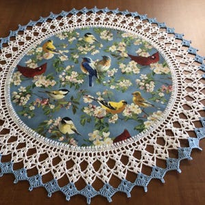 May include: Round decorative doily featuring a central fabric panel with a blue background and a pattern of birds and flowers. The doily has a white and light blue crocheted lace border. A charming home decor item.