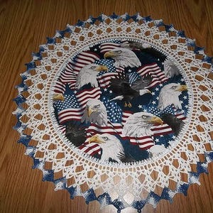 May include: Round patriotic doily featuring a central design of American flags and bald eagles. The doily has a white crocheted border with blue accents. The design is centered on a wooden surface.
