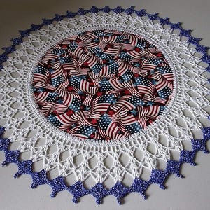 May include: A round, patriotic doily featuring a central fabric circle patterned with American flags. The doily has a white, open-weave crochet design with a scalloped edge trimmed in blue glitter.