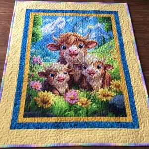 May include: A rectangular quilt featuring a central printed panel of three cartoon highland cows in a grassy field with colorful flowers. The panel is framed by a dark blue border and an outer yellow border with a subtle, wavy quilted texture.