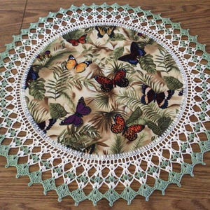 May include: Round decorative placemat featuring a butterfly and tropical leaf pattern. The center fabric is surrounded by a white crocheted border with a light green scalloped edge. The butterflies are in shades of orange, purple, and blue.