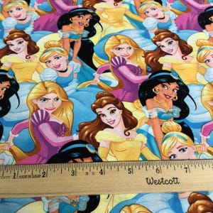 May include: Fabric featuring a repeating pattern of animated princess characters in various poses. The princesses are depicted in their signature gowns, with a ruler in the foreground. The fabric has a light blue background.