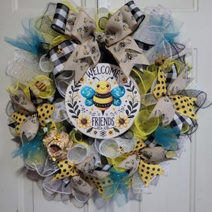May include: A decorative wreath with a central sign displaying a cartoon bee and the words "WELCOME FRIENDS." The wreath incorporates yellow, black, and white mesh, patterned ribbons, and bee-themed embellishments. A charming home decor item.
