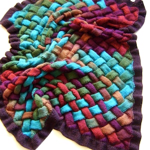 Könnte beinhalten: Ein farbenfroher gestrickter Schal mit einem gewebten Muster in den Farben Türkis, Lila, Braun und Grün. Der Schal hat einen gewellten Rand und ist aus einem weichen, strukturierten Garn gefertigt.