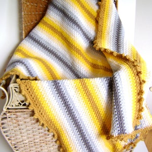 May include: A handmade crochet blanket with a striped pattern in yellow, gray, white, and brown. The blanket has a scalloped edge and is draped over a wicker chair. The blanket is a cozy home decor item.