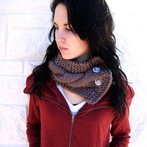 May include: A brown knitted cowl scarf with two silver buttons. The scarf is wrapped around the neck of a person wearing a red zip-up hoodie over a white shirt. The background is a white brick wall.