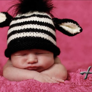 May include: A sleeping baby wearing a black and white striped knit hat with attached ears and a black pom-pom. The baby is resting on a pink textured blanket against a pink background.