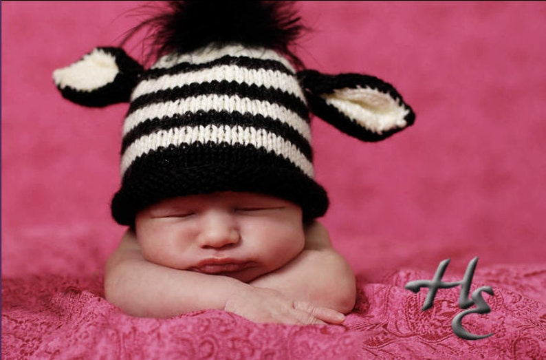 May include: A sleeping baby wearing a black and white striped knit hat with attached ears and a black pom-pom. The baby is resting on a pink textured blanket against a pink background.