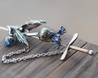 May include: A silver brooch depicting a thistle and a small sword. The thistle has blue and green detailing, and the sword has a gold-coloured handle. A silver chain links the two elements together.