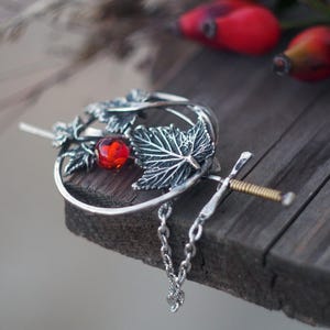 May include: A silver hair pin with a decorative design featuring a red gemstone, leaves, and a chain. The pin is set against a blurred background with red berries and foliage.