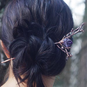 Könnte beinhalten: Eine kupferfarbene Haarnadel mit einem astähnlichen Design und einem großen, facettierten lila Kristall. Das Haaraccessoire ist in dunklem Haar gesetzt, mit einer zweiten, einfachen Kupfernadel sichtbar. Der Hintergrund ist unscharf.