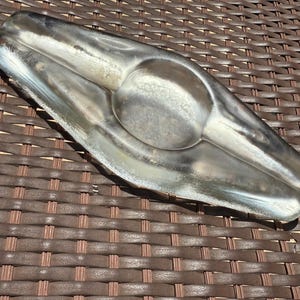 Puede incluir: Un cenicero plateado con forma redondeada y alargada. El cenicero tiene una hendidura circular central y un diseño curvo. La superficie metálica refleja la luz, resaltando su textura lisa. El cenicero está diseñado para cigarrillos o puros.