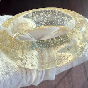 May include: A clear, resin bangle bracelet with gold leaf flakes suspended within. The bracelet has a rounded, smooth shape and is approximately 2.5 inches in diameter. The gold flakes add a touch of sparkle and luxury to the accessory.