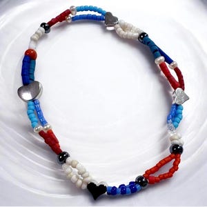 Puede incluir: Un collar de cuentas con una combinación de colores patrióticos. El collar presenta cuentas rojas, blancas y azules, con dijes plateados en forma de corazón y un dije negro en forma de corazón. Las cuentas están ensartadas en un diseño en capas.
