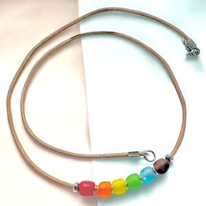 May include: A necklace featuring a tan leather cord with a silver-toned metal clasp. The cord is accented with a row of six small, irregularly shaped beads in red, orange, yellow, green, light blue, and dark brown, separated by silver spacers.