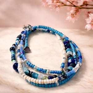 Puede incluir: Una pulsera de múltiples hilos con cuentas azules y blancas. La pulsera incluye un pequeño dije de corazón plateado y un cierre plateado. Las cuentas varían en tamaño y textura, creando un efecto en capas. Un accesorio elegante.