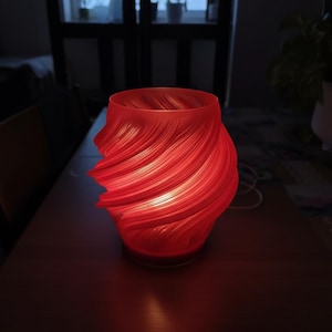 Può includere: Lampada decorativa rossa con un design strutturato e a spirale. La lampada ha una parte superiore ampia e una base arrotondata. La fonte di luce è all'interno, creando un bagliore caldo. La lampada è su una superficie di legno scuro. Altezza circa 20 cm.