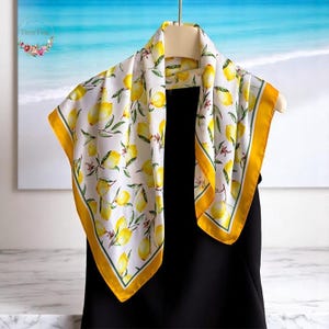 May include: A white silk scarf with a yellow lemon print and a yellow border. The scarf is folded and draped over a black dress.