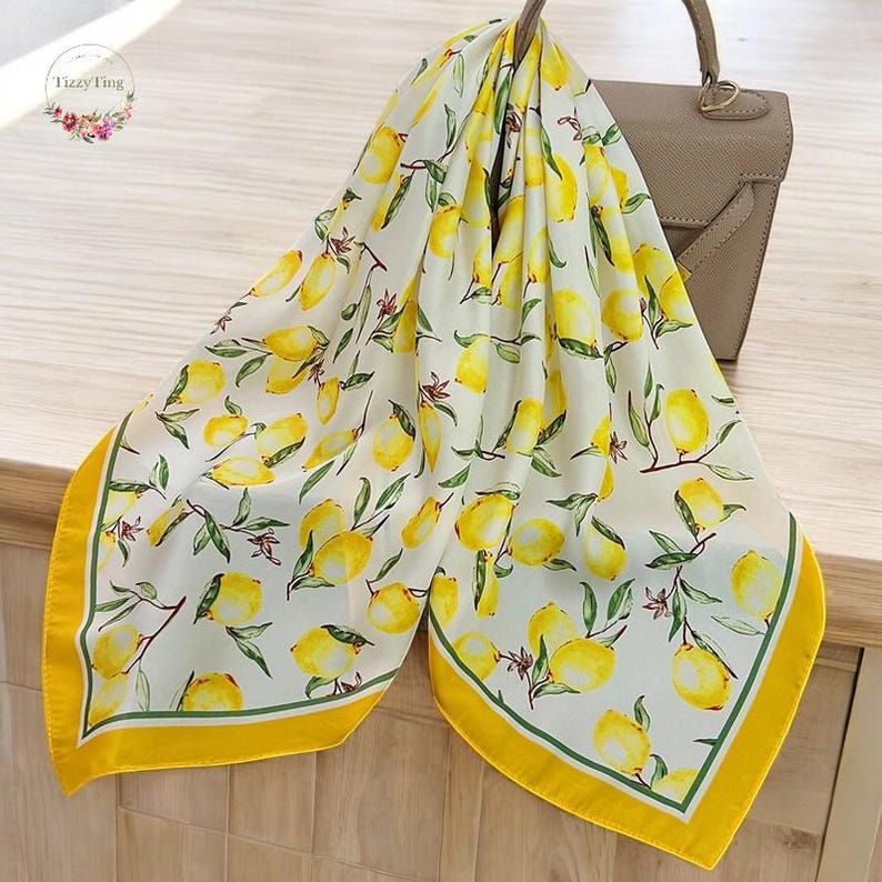 May include: A white silk scarf with a lemon pattern. The scarf features yellow lemons with green leaves and stems. The border is yellow with a green trim. The scarf is draped over a tan handbag.