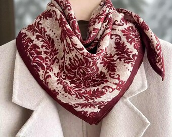 May include: A square scarf featuring a burgundy and cream floral design. The scarf is displayed over a white coat, highlighting the detailed pattern and colour contrast. The edges of the scarf are a darker burgundy.