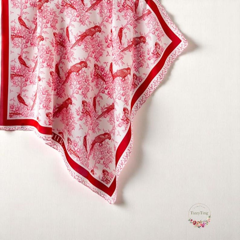 May include: A white silk scarf with a red border and a repeating pattern of red parrots and apples. The scarf is folded in a triangular shape.
