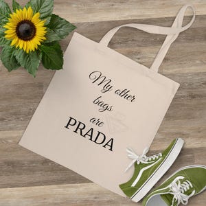 Peut inclure: Sac fourre-tout crème avec le texte "My other bags are PRADA" en écriture noire. Le sac a de longues anses et se trouve à côté d'une paire de baskets vertes et d'un tournesol. Le sac est en toile.