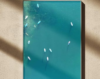 May include: A framed print featuring an aerial view of a turquoise body of water with numerous white paddleboards. The artwork is set against a neutral-toned background, with shadows cast across the surface.