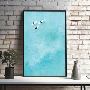 Op de afbeelding: Een luchtfoto van drie mensen op surfplanken in een turquoise blauwe oceaan. Het water is kalm en de lucht is helder.