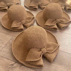 May include: A collection of tan crochet sun hats, each featuring a rounded crown, a wide brim, and a prominent decorative bow attached to the front. The hats are crafted from a textured, woven material in a consistent light brown color throughout.