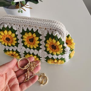 May include: A cream-colored crochet clutch featuring a sunflower pattern in yellow, green, and brown. The bag has a gold chain strap and clasp. The clutch is handmade and has a soft, textured appearance.