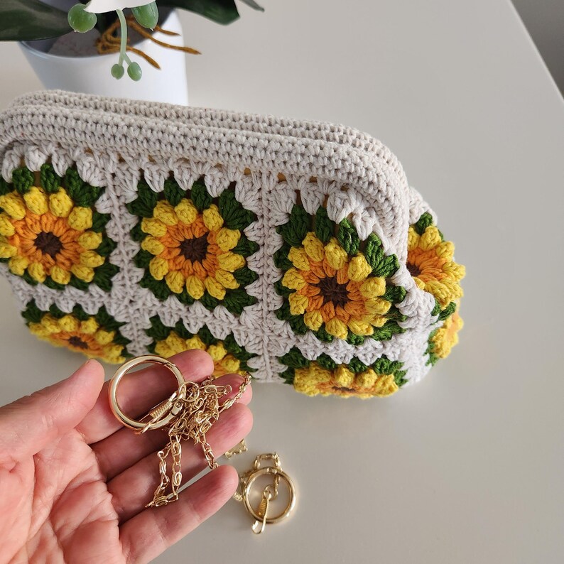 May include: A cream-colored crochet clutch featuring a sunflower pattern in yellow, green, and brown. The bag has a gold chain strap and clasp. The clutch is handmade and has a soft, textured appearance.