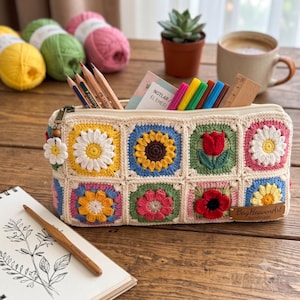 Könnte beinhalten: Ein rechteckiges Häkel-Etui mit einem Gitter aus bunten floralen Granny Squares in Gelb-, Blau-, Grün-, Rosa- und Rottönen. Das Etui verfügt über einen Reißverschluss mit einem kleinen Gänseblümchen-Anhänger und einem braunen Lederetikett.