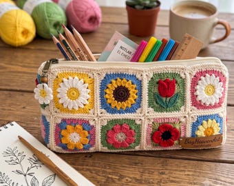 May include: A rectangular crochet pencil case featuring colorful granny squares with floral motifs including daisies, sunflowers, and tulips. The case has a zipper closure with a small daisy charm and a leather tag labeled BagHeavenArt.