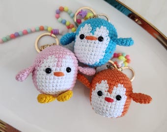 May include: Three handmade crochet penguin keychains in pink, blue, and orange. Each penguin has a white belly, black eyes, and an orange beak. The keychains have gold-colored rings and colorful beaded straps.