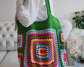 May include: A large tote bag crafted from green yarn, featuring a prominent central square motif composed of concentric rows of multi-colored crochet stitches in shades of pink, yellow, orange, purple, and blue, with a single shoulder strap.