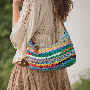 May include: A hobo-style shoulder bag crafted from multicolored crocheted yarn featuring horizontal stripes in shades of blue, green, yellow, red, white, and black, complemented by a brown leather strap and silver-toned metal hardware.